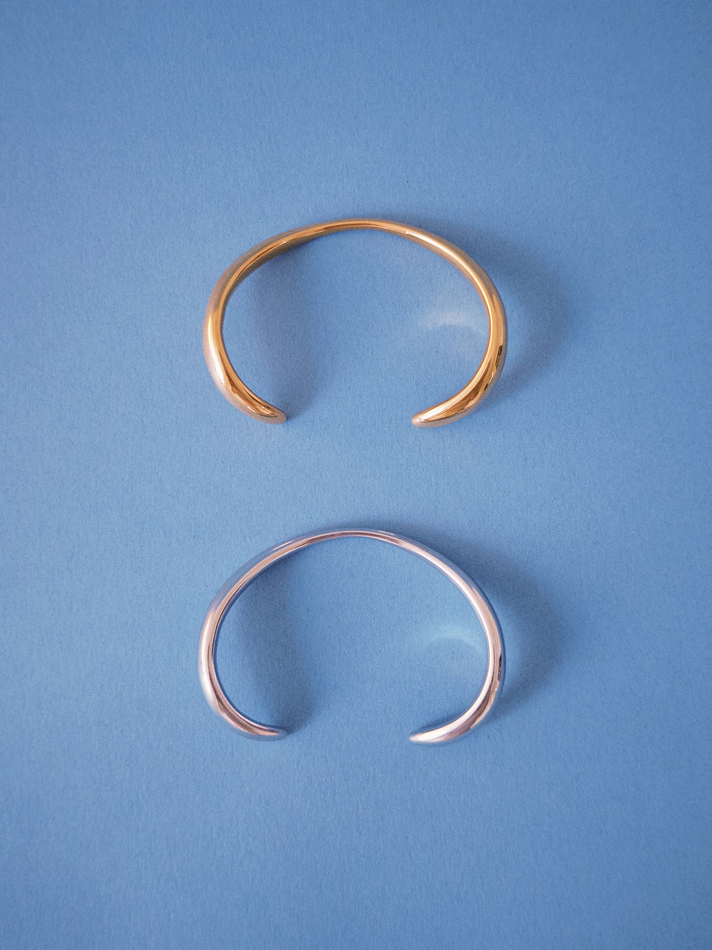 Minimal open cuff bracelets in gold and silver finishes on blue background