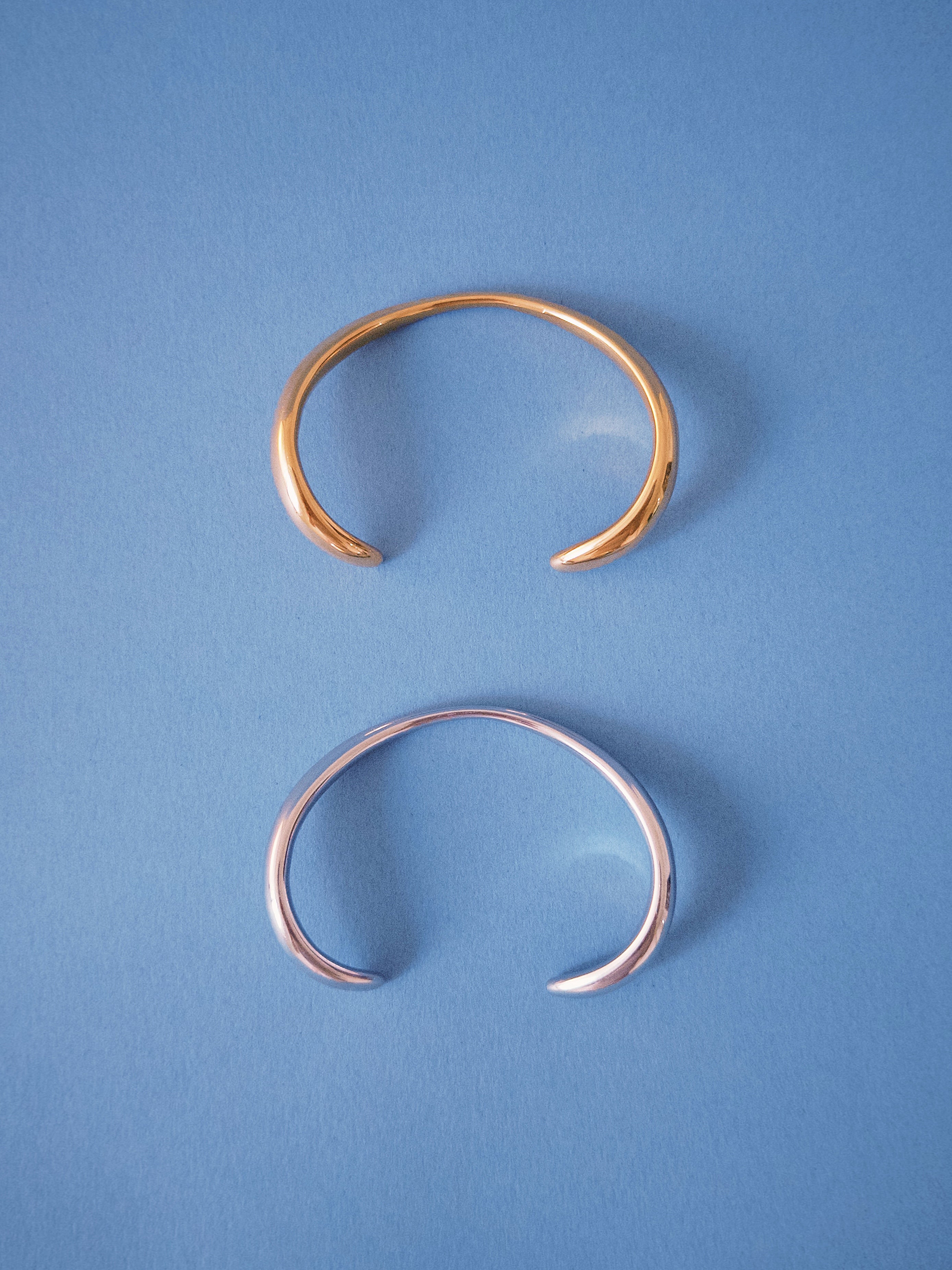 Minimal open cuff bracelets in gold and silver finishes on blue background